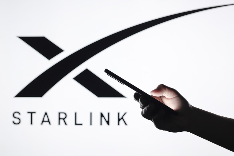 In this photo illustration, the Starlink logo seen in the background with a silhouette hand holding a smartphone.