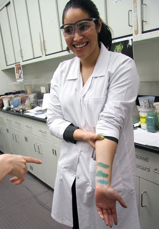 christine mielke aka temptalia wearing a white lab coat and lab glasses with green makeup swatches on her arm