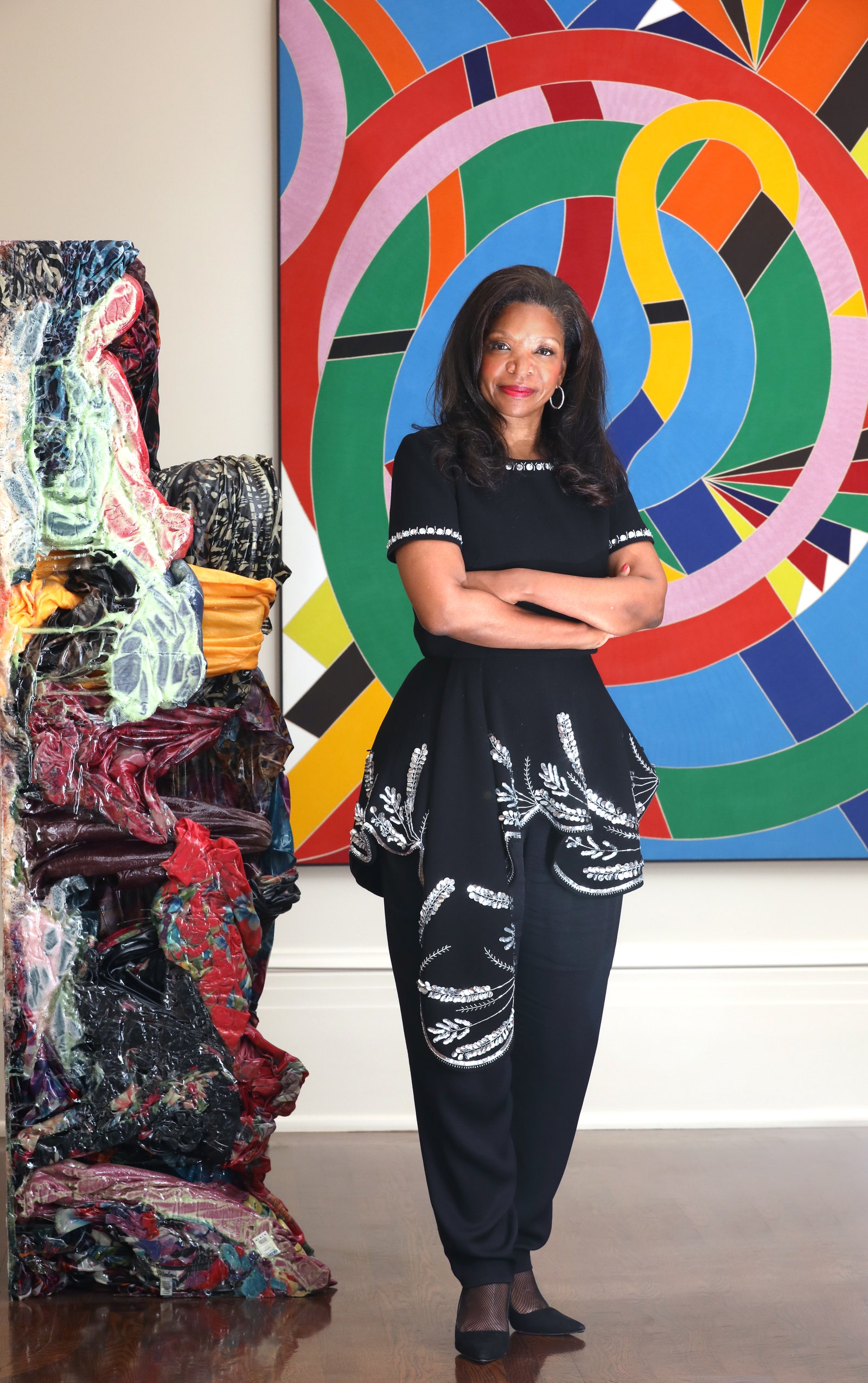 Pamela Joyner standing in front of one artwork on canvas hanging on white wall to left a multicolored sculptural piece