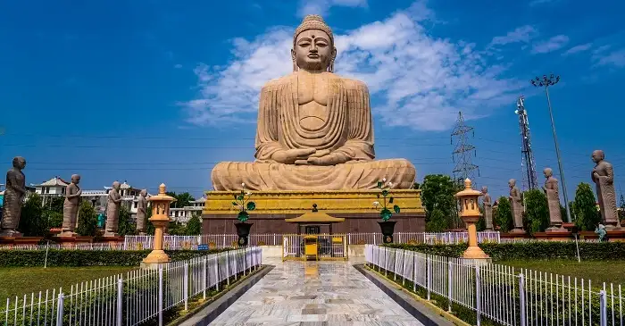 A large stone statue of a seated Buddha surrounded by smaller statues and greenery, set against a blue sky with scattered clouds in Bodh Gaya.