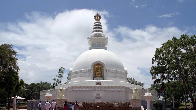 White pagoda in Rajgir surrounded by lush trees and people, Vishwa Shanti Stupa symbolizes world peace with golden statues depicting Buddhas life stages.