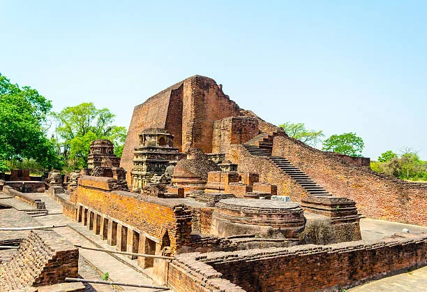 Ruins of an ancient brick structure with multiple levels, stairs, and stone carvings, reminiscent of the Buddhist monastic university of Nalanda, surrounded by trees under a clear blue sky.