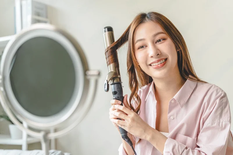 A women curling her hair with a hair curler looking in the mirror. 