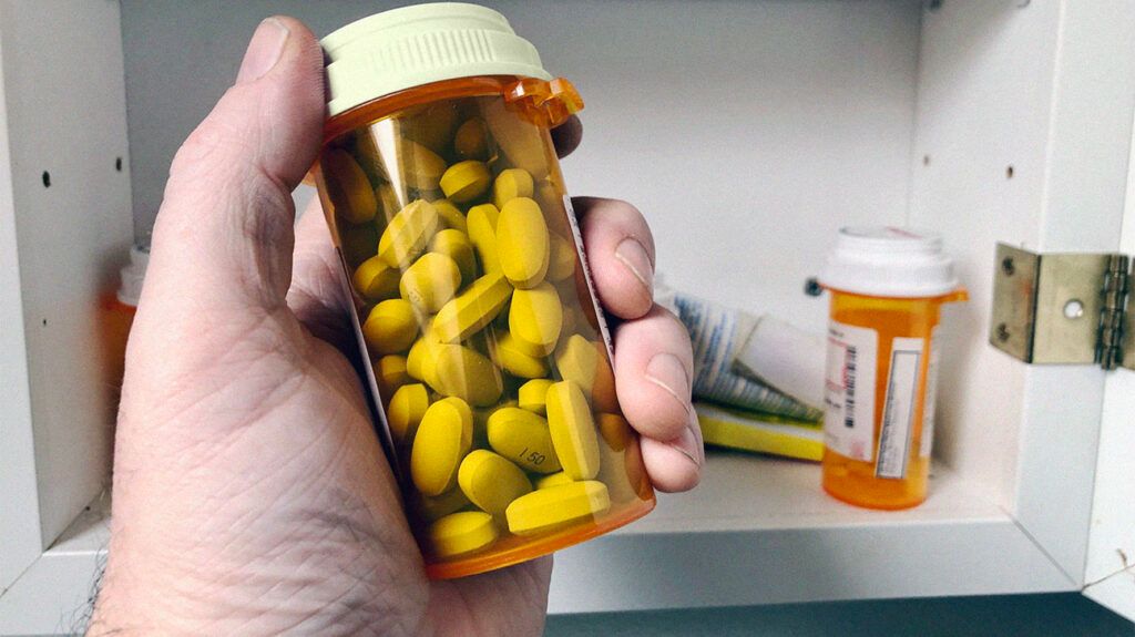 Man's hand holding bottle of anticholinergic drugs with a medicine cabinet in the background.