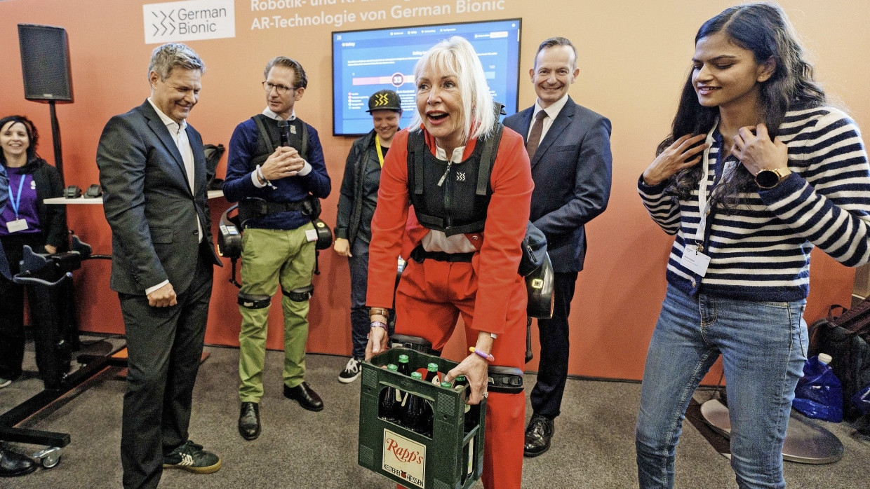 Sie packt es an: Hessens Digitalministerin Kristina Sinemus hebt unter den Augen von Bundeswirtschaftsminister Robert Habeck (links) und Bundesdigitalminister Volker Wissing (rechts hinter Sinemus) mit Exoskelett einen Kasten Ebbelwei.