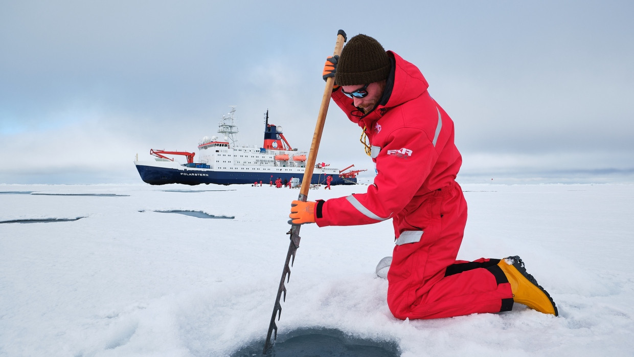 AWI-Ingenieur Daniel Scholz sägt ein Loch durch die Eisscholle, um ein Messinstrument ins Wasser zu lassen. Im Hintergrund ist die Polarstern zu sehen.