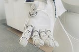 A white robot extends its hand to the viewer, many-jointed fingers outstretched in preparation to grasp something, perhaps mind-numbing piles of paperwork