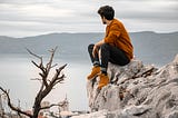 A young man in a burnt orange shirt and burnt orange shoes sits on a rock overlooking a lake