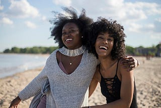 Two female friends embrace while smiling and walking along the beach