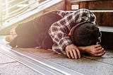 Homeless man curled up sleeping on pathway with bare feet and no blankets