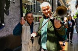 Two men standing on the sidewalk in downtown Belfast. Author’s husband is playing a street musician’s homemade string instrument similar to a violin