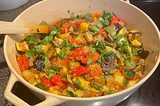 A large Dutch oven filled with summer seasonal vegetables topped with fresh basil. A wooden spoon is off to the side, ready to stir.