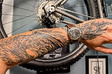 Closeup of the author’s forearm and hand with a black and grey tattoo of a firebird. He’s wearing a windup watch with a leather band and there’a bicycle wheel in the background.