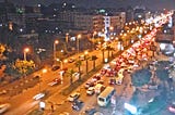 Nighttime heavy traffic on a wide 8-lane two-way road. Buildings are on both sides of the street. There are palm trees on the boulevard between traffic.