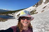 The author is very sunburnt, wearing a tie-dye hat and standing in front of a beautiful partially ice-covered lake on the side of a snowy mountain at St Mary’s Glacier in Colorado. The sky is blue without a cloud in it. There are more mountains and many trees in the background.