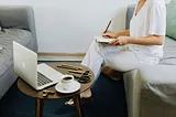 woman sits on a couch writing something in a notebook with a laptop and coffee on a small table in front of her.