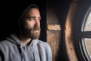 A bearded man wearing a hat looks out the window with a sad expression on his face