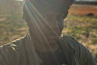 Black trans human standing in a field with the sun on their back. the human has a smirk, as if they know something the reader doesn’t.