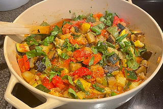 A large Dutch oven filled with summer seasonal vegetables topped with fresh basil. A wooden spoon is off to the side, ready to stir.