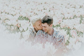 A man kisses the neck of another man as they sit in a field of flowers