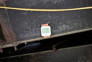 The image shows a small digital thermometer and humidity  device with a square shape, mounted on a rusted metal surface. The device displays temperature and humidity readings, with the top number showing “72.9” and the bottom number displaying “63%.” Above the device, a yellow cable or wire is loosely positioned. The surrounding area has a weathered, industrial appearance, with rust and signs of wear visible on the metal.