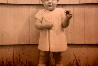 A young toddler standing outside a house and holding a round metal circle in her hand.