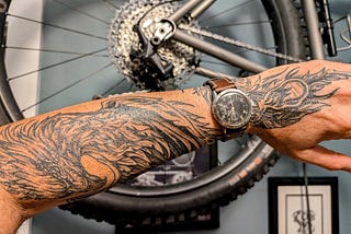 Closeup of the author’s forearm and hand with a black and grey tattoo of a firebird. He’s wearing a windup watch with a leather band and there’a bicycle wheel in the background.