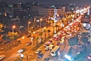 Nighttime heavy traffic on a wide 8-lane two-way road. Buildings are on both sides of the street. There are palm trees on the boulevard between traffic.