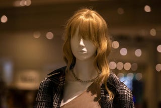 A mannequin wearing a blonde wig with a chain around its neck, dressed in a plaid jacket and undershirt