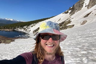 The author is very sunburnt, wearing a tie-dye hat and standing in front of a beautiful partially ice-covered lake on the side of a snowy mountain at St Mary’s Glacier in Colorado. The sky is blue without a cloud in it. There are more mountains and many trees in the background.