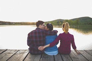 A couple kissing while the man (far left) hold hands with a second woman (right).