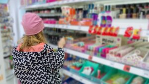 A girl with her back to the camera stands in a store, facing shelves of health and beauty products on display for sale. The girl appears to be the size of a tween or young teen and is wearing a black and white patterned coat and a pink winter cap.