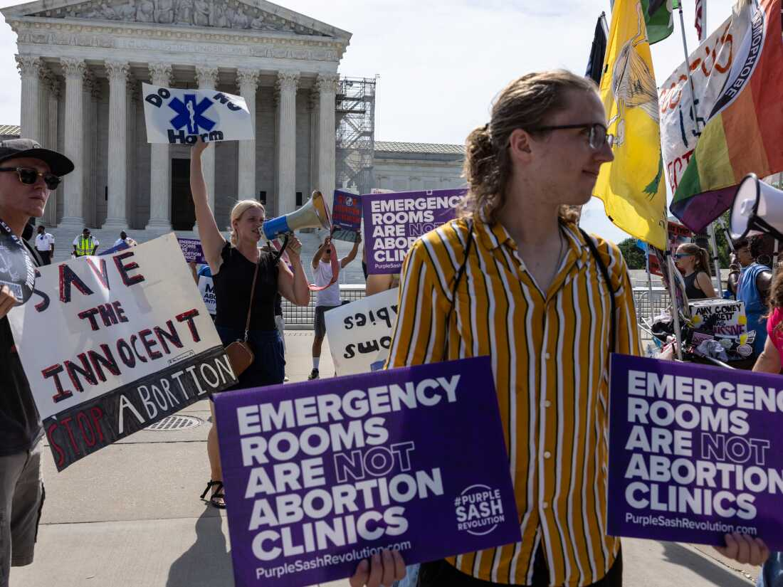 A young man in a striped yellow shirt holds two purple signs that both read, 