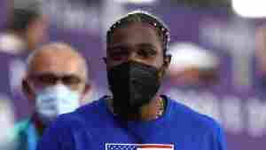 Olympic sprinter Noah Lyles wears a black KN95 mask and a blue t-shirt with an American flag on it. 