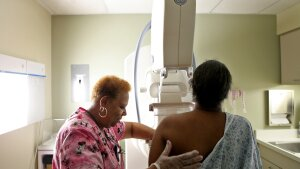 An African American woman is shown getting a mammogram. She is photographed from behind, so we see the back of her head and body as she stands facing a large X-ray machine. A health care professional wearing pink scrubs positions the woman in the machine.