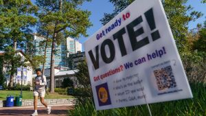 In the foreground, a sign encourages people to vote. In the background are buildings, trees and a young man walking.