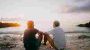 A photograph of a senior father and his adult son photographed from behind. They are sitting on the sand at the beach and looking out at the ocean at sunset while having a deep conversation.