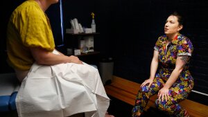 Dr. Stephanie Arnold, who is wearing a brightly colored jumpsuit, speaks with a patient who is sitting on an exam table with a medical drape over her lap.