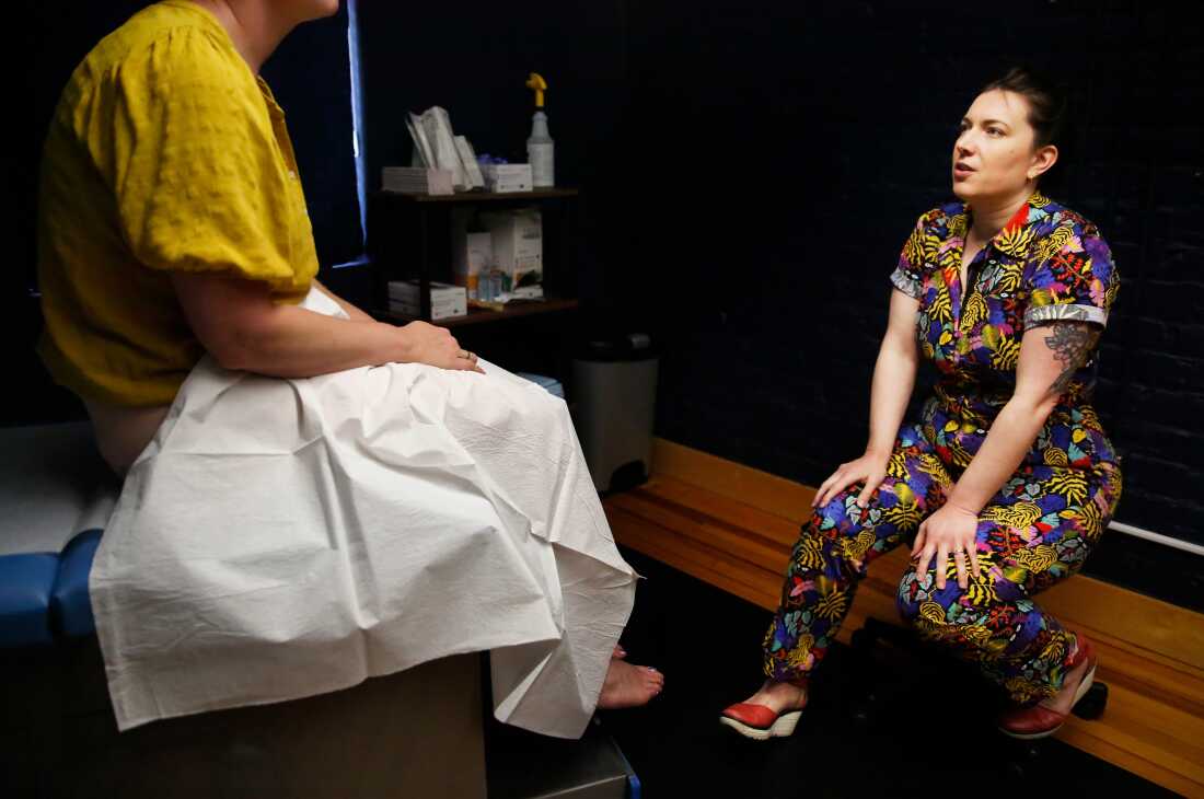 Dr. Stephanie Arnold, who is wearing a brightly colored jumpsuit, speaks with a patient who is sitting on an exam table with a medical drape over her lap.