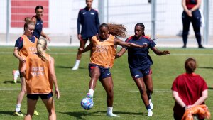 Several women from the U.S. Women's National Team practice soccer on a green soccer field. A few are wearing orange vests that say 