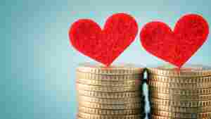 Two red felt hearts balance atop two separate stacks of gold coins against a light blue backdrop, representing people in a romantic relationship merging finances.