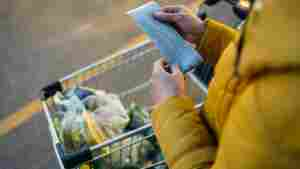 A high-angle, close-up photograph shows a person holding and reviewing a grocery store receipt in the parking lot, and their grocery cart full of purchased food sits in the background. 