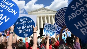 The image shows a bright blue sky and fluffy clouds above the Supreme Court building in the background, and protestors holding blue signs with white type that read, 