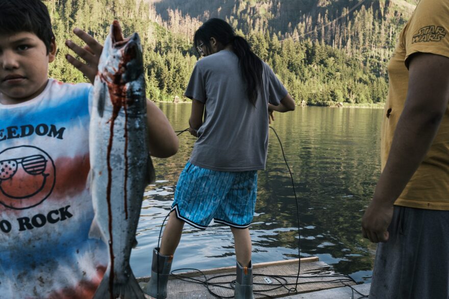 A person reeling in a fish as someone in a yellow shirt watches and a child holds up a fish off to the corner