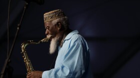 A Black man with a long white beard plays saxophone wearing a light blue robe and embroidered hat.