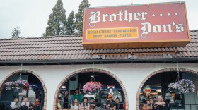 A group of people are smiling outside a patio restaurant with hanging flower pots and a sign on the roof that reads, "Brother Don's."
