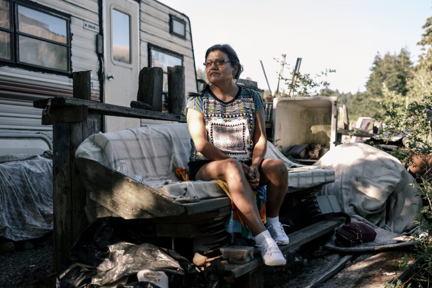 A woman sits outside of a mobile camper on a make-shift outdoor seating area surrounded by older weathered items.