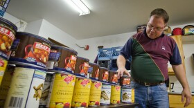 A man in jeans and a maroon, navy, and deep green polo shirt - straightens a collection of cans on a table as a collection of milk cartons are stacked behind him. 