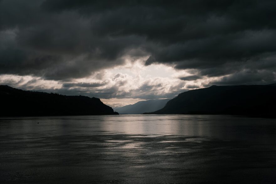 The sun breaks through dark clouds between an opening between mountains over a lake.