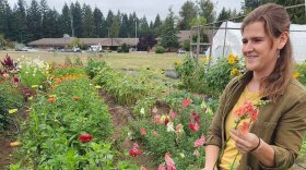 A woman wearing a brown and yellow shirt underneath stands in front of a row of flowers while holding some of the flowers in her hands.